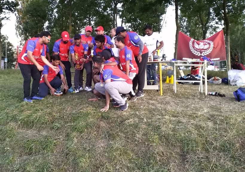 Players in red kit gathered under trees at the edge of the ground