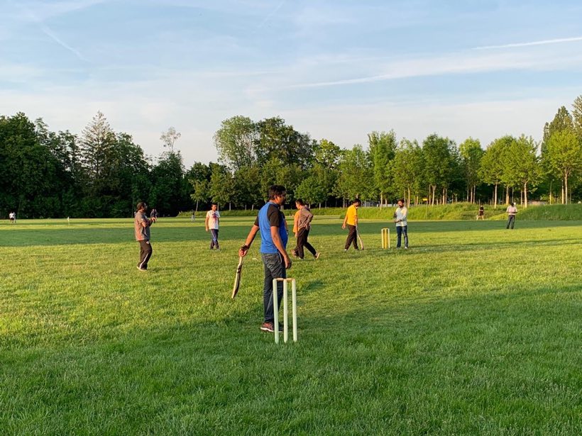 An evening match in progress on a grass outfield, trees along the boundary